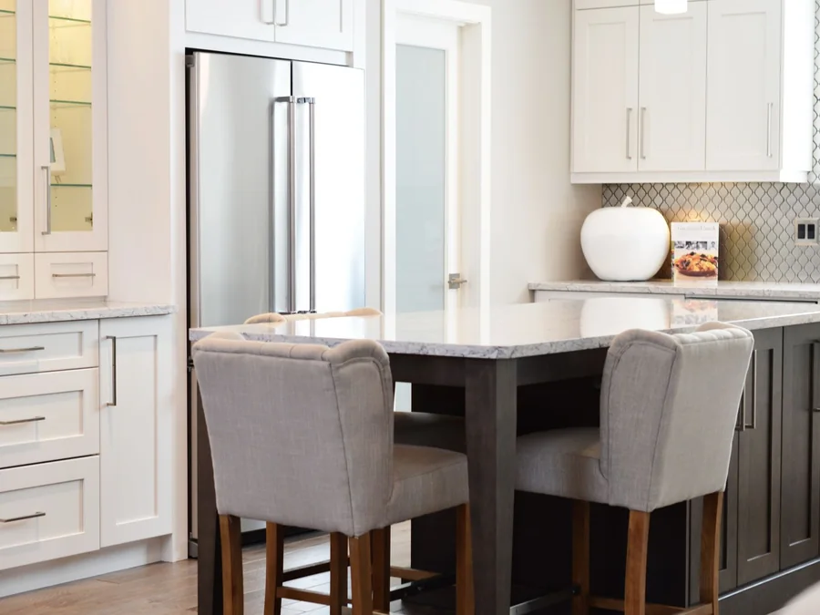 White kitchen with a stone island, upholstered stools and stainless steel refrigerator